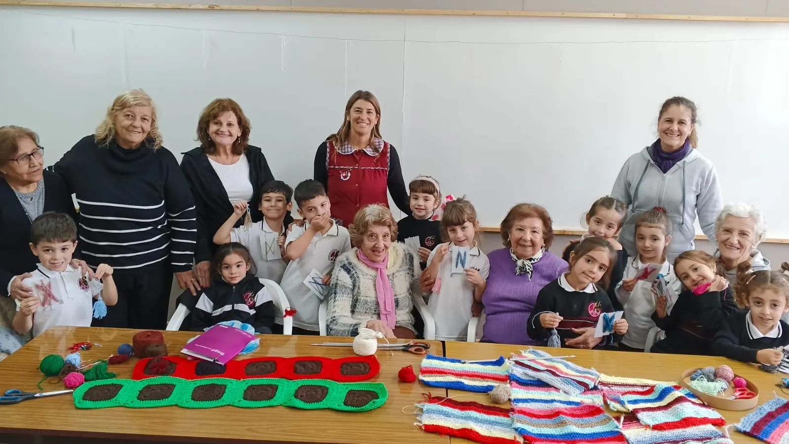Chicas y chicos del Nivel Inicial junto a un grupo de señoras mayores y sus maestras en un taller de tejido, con las labores terminadas sobre la mesa.